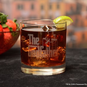 May include: A clear glass tumbler with the words "The Godfather" etched on the side. The glass is filled with a brown liquid and ice cubes. A lime wedge is on the rim of the glass.