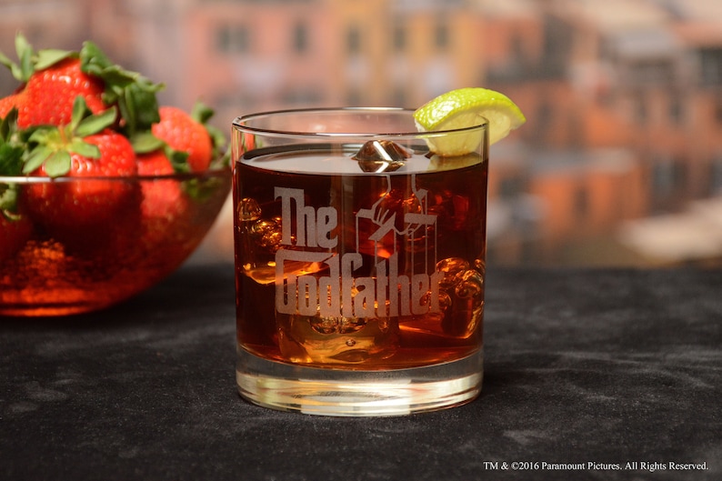 May include: A clear glass tumbler with the words "The Godfather" etched on the side. The glass is filled with a brown liquid and ice cubes. A lime wedge is on the rim of the glass.