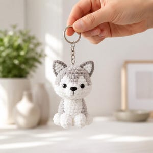 Może przedstawiać: Ręcznie robiony brelok do kluczy z psem husky wykonany na szydełku. Brelok przedstawia małego, szarego i białego psa husky z czarnymi oczami i czarnym nosem. Pies jest przymocowany do srebrnego kółka i łańcuszka.