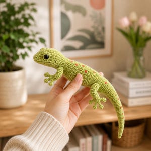 May include: A crocheted green lizard with red spots is held in a hand. The handmade reptile has detailed feet, a long tail, and black eyes. The lizard is a unique, handcrafted item.