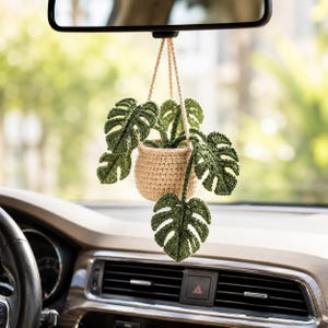 Puede incluir: Una planta colgante de ganchillo con hojas verdes y una maceta beige, suspendida del espejo retrovisor de un coche. La planta presenta patrones de hojas detallados, añadiendo un toque de naturaleza al interior del coche. El salpicadero del coche es visible en primer plano.