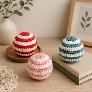 May include: Three handmade crochet balls in red and white, pink and white, and teal and white stripes. The balls are on a wooden surface with a framed picture and a vase with greenery in the background.