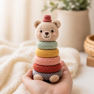 May include: A crochet bear stacking toy featuring a beige bear head with a small pink hat on top. The body consists of five stacked rings in light green, yellow, light pink, dark red, and grey, with two small beige feet at the base of the toy.
