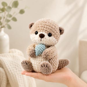 Op de afbeelding: Een handgemaakte gehaakte otter knuffel in bruin, crème en blauw. De otter heeft een ronde kop, kleine oren en houdt een blauw hartvormig object vast. Het speelgoed is zacht en knuffelig, perfect voor een kinderkamer of als decoratief item.