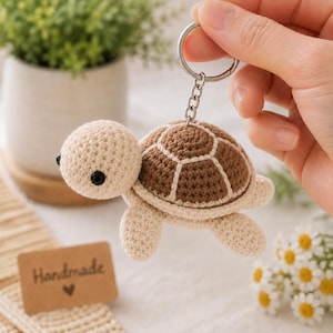 May include: A handmade crochet turtle keychain in beige and brown. The turtle has a beige head and limbs, a brown shell with beige accents, and black eyes. The keychain is attached to a silver metal ring. A small tag reads "Handmade".
