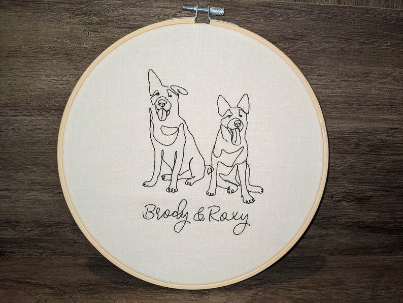 Op de afbeelding: Borduurwerk op een hoepel met twee honden, Brody en Roxy, in een eenvoudige lijntekening stijl.
