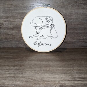 Op de afbeelding: Borduurwerk op een hoepel met een lijntekening van twee honden, een op de ander, met de tekst "Lady & Kevin" eronder.