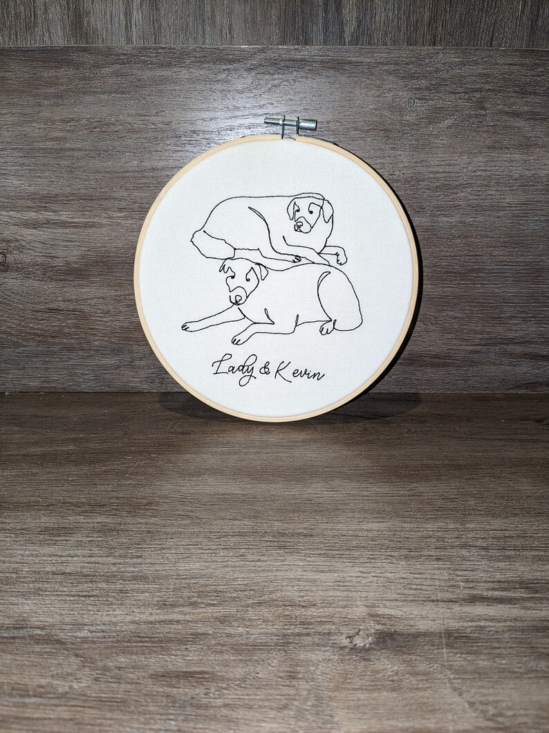 Op de afbeelding: Borduurwerk op een hoepel met een lijntekening van twee honden, een op de ander, met de tekst "Lady & Kevin" eronder.