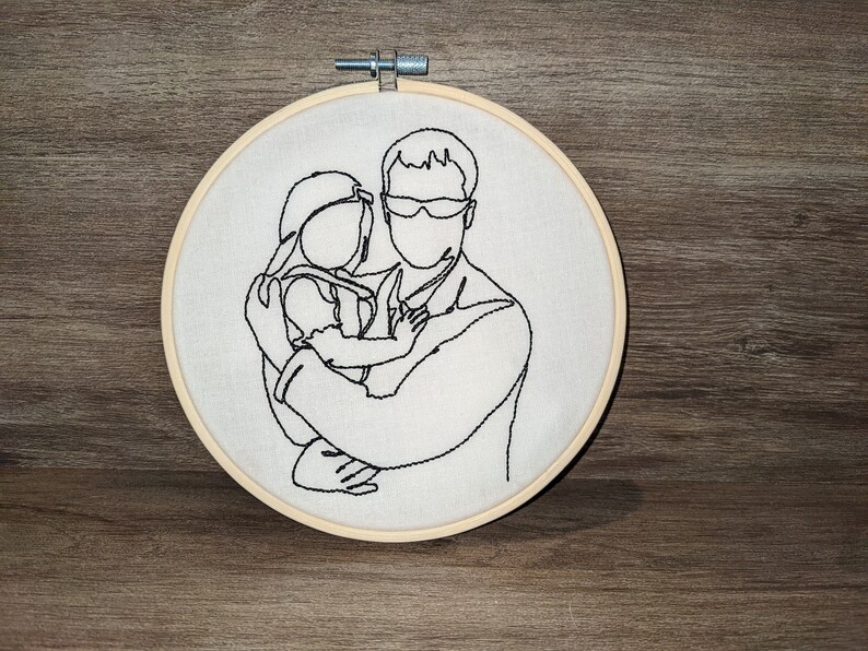 Op de afbeelding: Borduurwerk op een borduurring met een lijntekening van een persoon die een kind vasthoudt. De tekening is in zwart garen op een witte achtergrond.