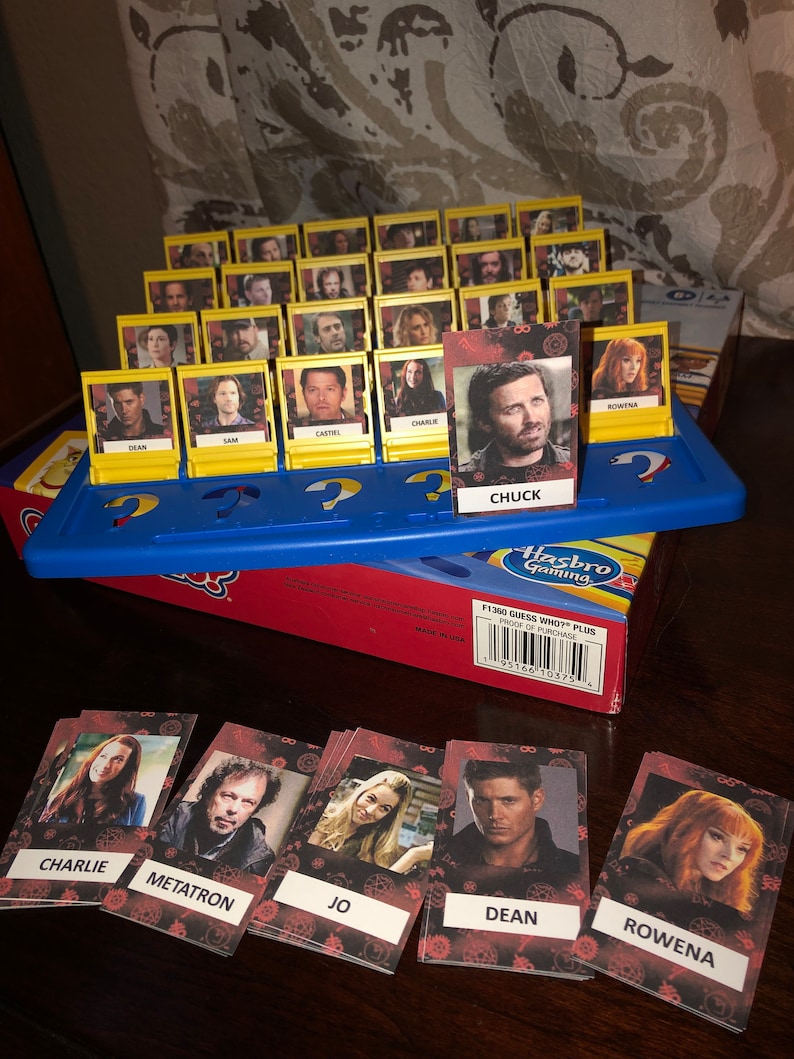 Puede incluir: Un tablero de juego Guess Who? con cartas de personajes que representan personajes del programa de televisi&oacute;n Supernatural. Las cartas son amarillas con un borde negro y muestran una foto de un personaje del programa. Las cartas est&aacute;n dispuestas en una cuadr&iacute;cula en el tablero. El tablero de juego es azul y tiene un logotipo de Hasbro Gaming rojo y blanco. El tablero de juego est&aacute; sobre una mesa de madera.
