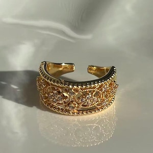 May include: A gold-colored adjustable ring with an ornate design. The ring features a detailed pattern with small, sparkling accents. The open design allows for size adjustment. The ring is set against a soft, white background.