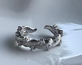 May include: A silver adjustable ring with a wave pattern. The ring has small, clear gemstones and an open design. The ring is displayed on a white surface.