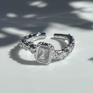 May include: A silver-toned open ring featuring a central rectangular clear gemstone. The band has a textured, organic surface with small raised bumps throughout, creating a rugged appearance. The ring is adjustable with a gap at the back.