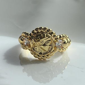 May include: A gold-colored ring featuring a central medallion with a profile design, surrounded by a beaded border. Two small, clear gemstones flank the medallion. The ring has a detailed, ornate design.