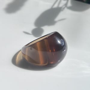 May include: A large, oval-shaped ring with a polished, translucent appearance. The ring features a blend of brown and amber tones, creating a marbled effect. The ring is set against a white surface, with shadows adding depth.