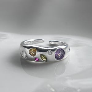 May include: A silver-colored ring with an open design, adorned with various colorful gemstones. The ring features a mix of round and oval-shaped stones in white, yellow, pink, green, and purple hues. The ring is set against a white background.