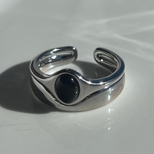 Peut inclure: Une bague ouverte de couleur argentée avec une finition lisse et polie, ornée d'une pierre noire ovale en son centre. La bague présente un design à anneau fendu avec une apparence incurvée et superposée, créant une forme fluide et continue.