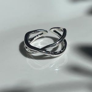 May include: A silver-colored ring with an open design. The ring features a crisscross pattern, with two bands of metal overlapping each other. The ring is set against a white background, with shadows in the background.
