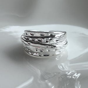 May include: A silver ring with a textured, layered design. The ring has a hammered appearance with small, evenly spaced indentations. The ring is set against a white background.