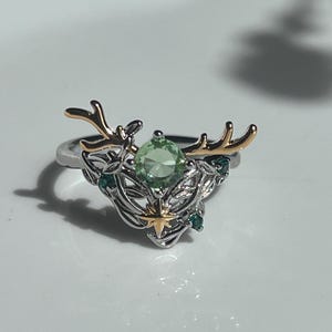 May include: A silver ring with gold-colored deer antlers and a light green gemstone center. The ring features intricate leaf and star details, with smaller green gemstones. The ring is set against a white background.