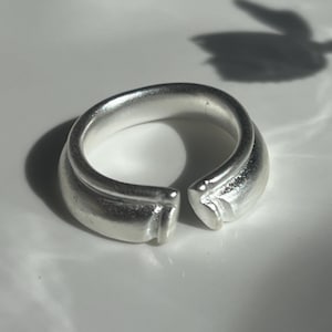 Könnte beinhalten: Ein silberfarbener offener Ring mit einem glatten, abgerundeten Band und matter Oberfläche. Der Ring weist an der Vorderseite eine deutliche Lücke auf, an der die beiden Enden aufeinandertreffen, was ein klares, durchgehendes Design ergibt.