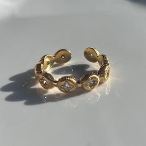 May include: A gold-colored adjustable ring with a repeating pattern of textured, circular elements. Each circular element is adorned with a small, square-shaped, clear gemstone. The ring is open at the top.