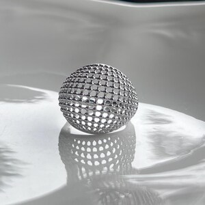 May include: A silver sphere with a lattice design, resting on a white surface. The sphere's surface is covered in evenly spaced, small, circular openings. The design creates a play of light and shadow, enhancing its three-dimensional form.