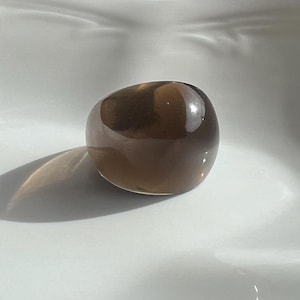 May include: A smooth, rounded, brown gemstone ring. The ring is a solid, opaque color with a polished surface. The ring is sitting on a white, curved surface, with a shadow cast to the left.