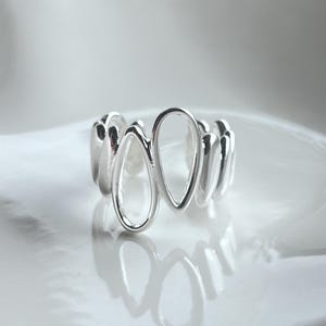 May include: A silver ring with an open, abstract design. The ring features a series of elongated, teardrop-shaped openings, creating a modern and minimalist aesthetic. The polished metal reflects light, highlighting the ring's smooth surface and unique form.