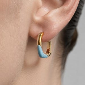 Könnte beinhalten: Ein goldener Creolen-Ohrring in rechteckiger Form mit glatter Oberfläche. Die untere Hälfte des Ohrrings ist mit hellblauem Emaille beschichtet, während die obere Hälfte aus poliertem Gold besteht. Der Ohrring wird mit einem Klappverschluss befestigt.