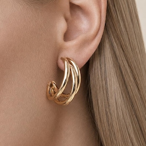 May include: A gold-toned hoop earring featuring a triple-layered design with three interconnected, smooth, polished circular bands that curve around the earlobe, creating a layered, multi-hoop appearance with a reflective metallic finish.