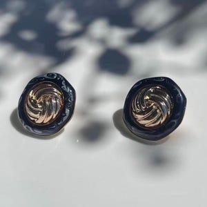 May include: A pair of gold-toned stud earrings. Each earring features a swirling gold center surrounded by a dark blue, patterned border. The earrings are set against a white surface, with shadows in the background.