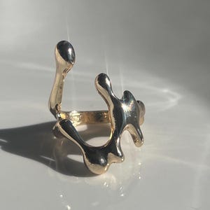 May include: A gold-coloured ring with an abstract, fluid design. The ring has a band and several organic, blob-like shapes that extend upwards. The ring is set against a white surface, with shadows adding depth.