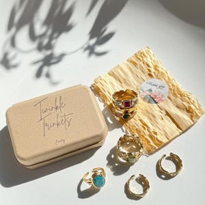 May include: A collection of gold-toned rings with various designs and gemstones, including a turquoise stone ring. The rings are displayed with a small, tan box labeled "Twinkle Trinkets" and a "Thank you" sticker on a bed of shredded paper.