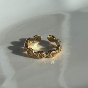 May include: A gold-colored adjustable ring featuring a series of oval-shaped, textured links. Each link has a central, clear stone. The ring is open at the top, allowing for size adjustment. The ring is on a white surface.