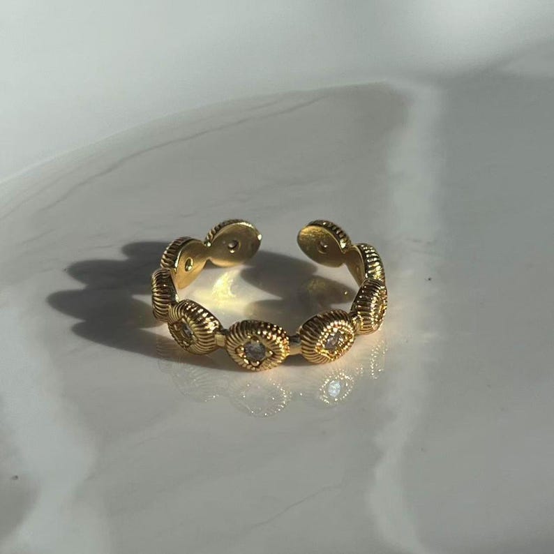 May include: A gold-colored adjustable ring featuring a series of oval-shaped, textured links. Each link has a central, clear stone. The ring is open at the top, allowing for size adjustment. The ring is on a white surface.
