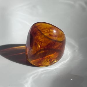 Op de afbeelding: Een dikke, doorschijnende ring met een gemarmerd patroon in warm amber en donkerbruin. Het gladde, gepolijste oppervlak van de ring vangt het licht, waardoor er diepe, wervelende kleurvariaties in het materiaal ontstaan.