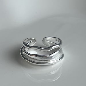 May include: A silver ring with a distinctive, layered design. It has a wavy upper band and a broader, textured lower band. The open design allows for adjustable sizing. The ring is made of a shiny, reflective metal.