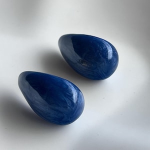 May include: A pair of smooth, teardrop-shaped earrings featuring a deep navy blue color with swirling white marbled patterns throughout the glossy, polished surface. These rounded, organic-shaped accessories have a reflective finish and solid structure.