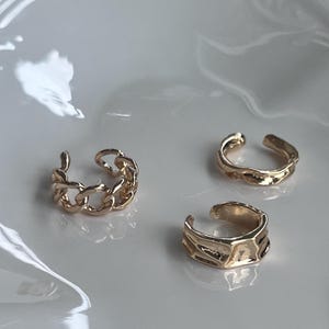Peut inclure: Trois ear cuffs dorées de différents modèles. L'un présente un motif de chaîne, tandis que les deux autres ont des surfaces texturées. Les ear cuffs sont présentés sur une surface blanche.