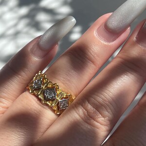 May include: A gold-toned ring with three diamond-shaped stones. The ring has an ornate, floral design with a textured finish. The ring is on a finger with long, light-colored nails.