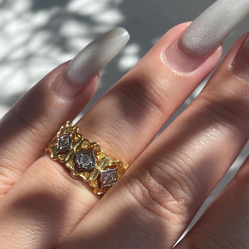 May include: A gold-toned ring with three diamond-shaped stones. The ring has an ornate, floral design with a textured finish. The ring is on a finger with long, light-colored nails.