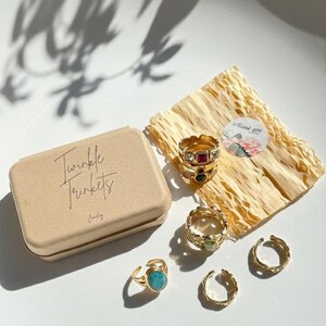 May include: An assortment of gold-toned rings with colorful gemstones and textures. A tan box with the words "Twinkle Trinkets" is next to the rings. A small sticker that says "Thank you" is on the right side of the image.