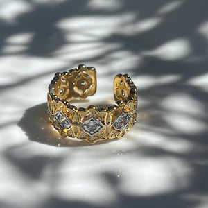 Op de afbeelding: Een goudkleurige verstelbare ring met een sierlijk ontwerp. De ring heeft een herhalend patroon van ruitvormige accenten met kleine, heldere stenen. De ring is aan de onderkant open, waardoor de maat kan worden aangepast. De ring staat tegen een witte achtergrond.