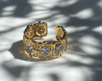 以下が含まれることがあります： 金色の装飾的なデザインの調節可能なリング。 リングは、小さな透明な石が付いた、ひし形アクセントの繰り返し模様が特徴です。 リングは下部が開いており、サイズ調整が可能です。 リングは白い背景に配置されています。