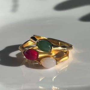Pu&ograve; includere: Tre anelli dorati con pietre preziose ovali verdi, rosse e bianche. Gli anelli sono impilati, con le pietre preziose rivolte verso l'alto. Gli anelli hanno un design strutturato e organico.