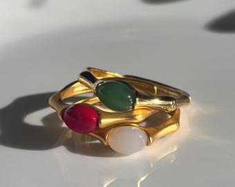 May include: Three gold-colored rings with oval-shaped gemstones in green, red, and white. The rings are stacked on top of each other, with the gemstones facing upwards. The rings have a textured, organic design.