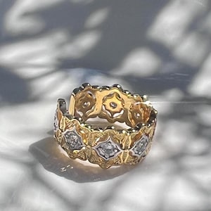May include: A gold ring with a repeating floral design, accented with small diamonds. The ring has a textured, openwork pattern and a bright, polished finish. The ring is likely a wedding band or eternity ring.