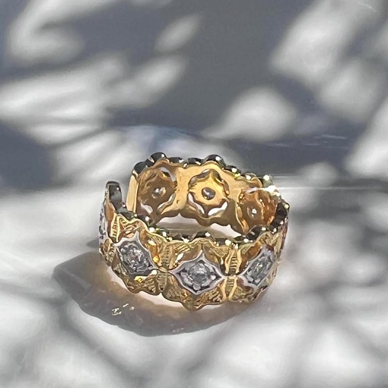 May include: A gold ring with a repeating floral design, accented with small diamonds. The ring has a textured, openwork pattern and a bright, polished finish. The ring is likely a wedding band or eternity ring.
