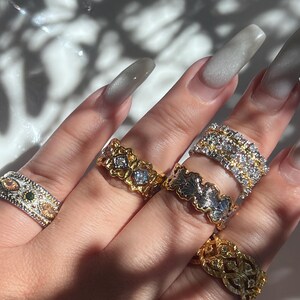 May include: A hand displaying several ornate rings. The rings feature various designs, including floral patterns, geometric shapes, and gemstone accents. The rings are gold and silver-toned, with clear and colored stones. The nails are long and painted a light gray color.